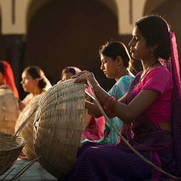 A group of women, including one in the foreground wearing a pink and purple sari, are seated and weaving large, round baskets from a light-colored material in an outdoor area. These items are handmade home decor items.