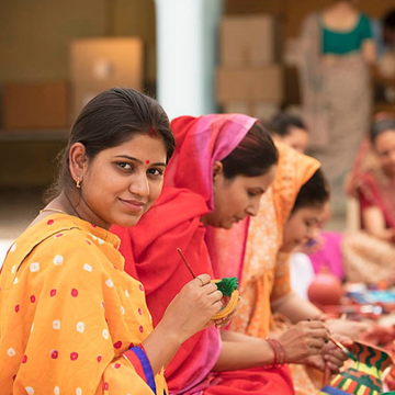 A group of women, seated on the floor in a brightly lit room, are engaged in intricate embroidery work. The woman in the foreground is smiling at the camera, holding a piece of fabric with a colorful floral design, which are handmade home decor items.