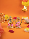 A pair of small, gold-plated idols of the Hindu deities, Lord Ganesha and Goddess Lakshmi, sitting side-by-side on lotus-style bases. The idols have pink-painted garments and are placed on a vibrant orange surface decorated with scattered rose petals and yellow and white marigold flowers. In the background, there is a golden-colored votive candle holder on the left and a bell-shaped decoration on the right.