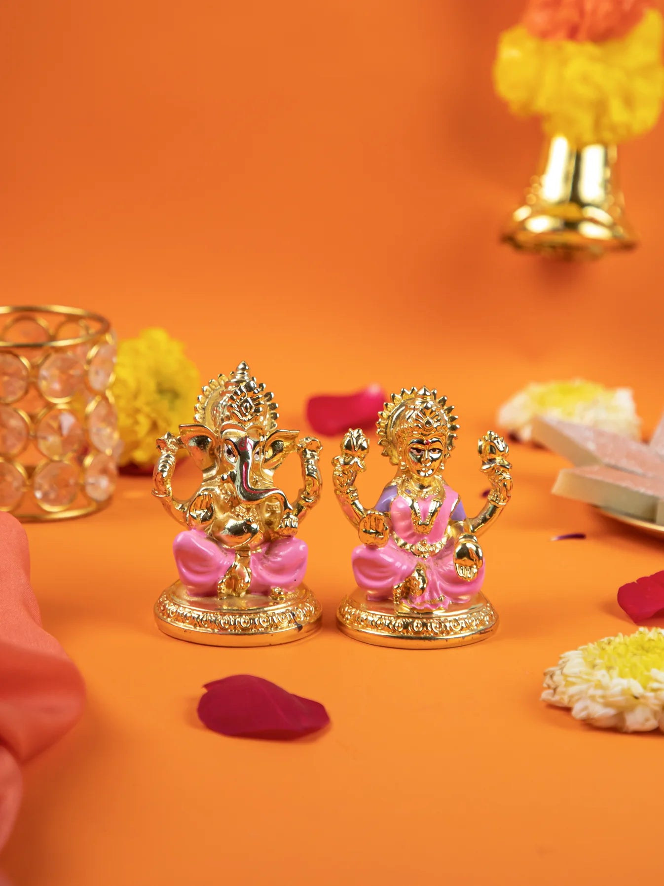 A pair of small, gold-plated idols of the Hindu deities, Lord Ganesha and Goddess Lakshmi, sitting side-by-side on lotus-style bases. The idols have pink-painted garments and are placed on a vibrant orange surface decorated with scattered rose petals and yellow and white marigold flowers. In the background, there is a golden-colored votive candle holder on the left and a bell-shaped decoration on the right.
