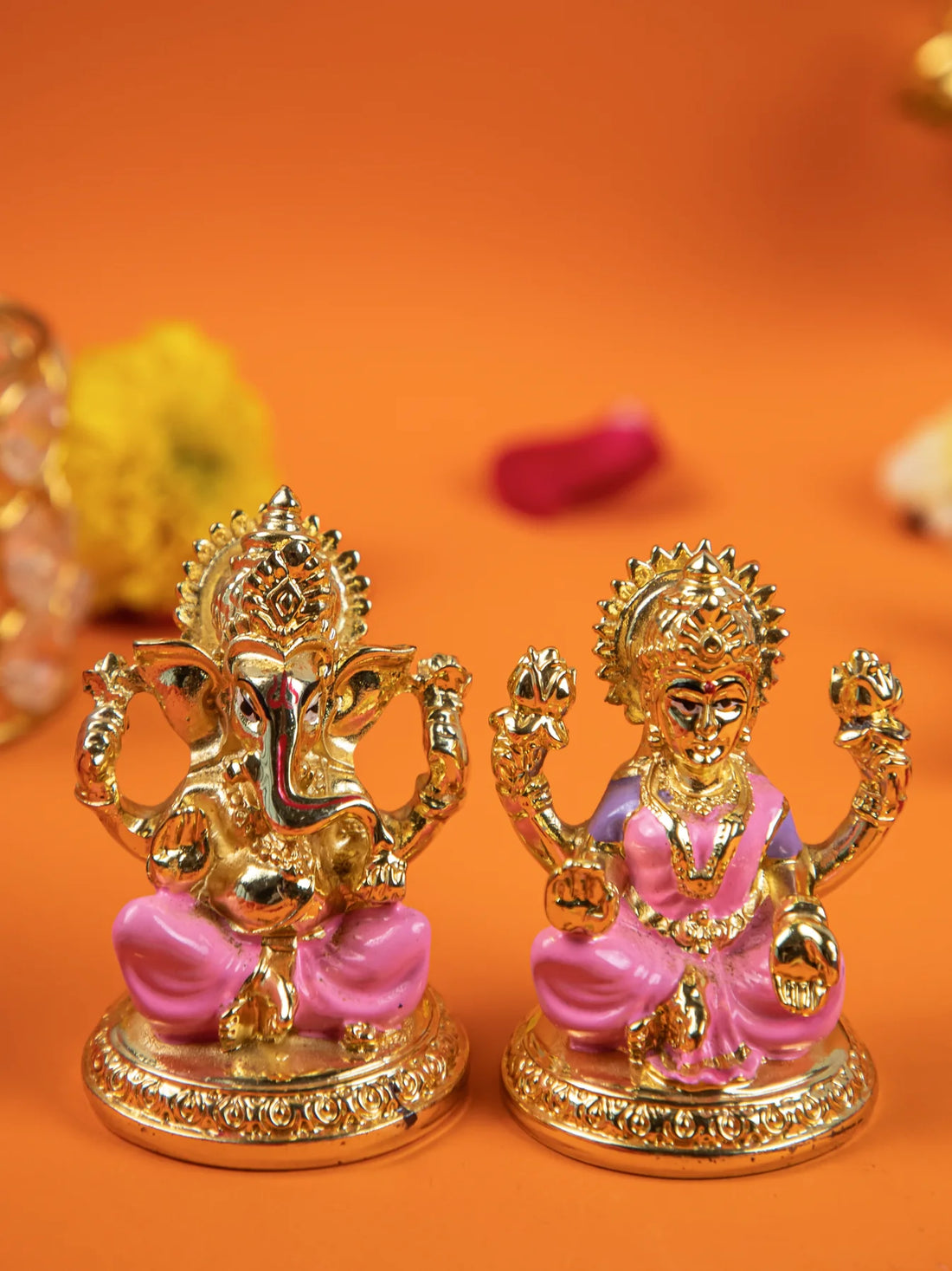 An image of an open teal blue velvet box containing two gold-colored idols of the Hindu deities Lakshmi and Ganesha with pink accents. The box is set on a peach-colored cloth, with yellow and white flowers and petals scattered around it.