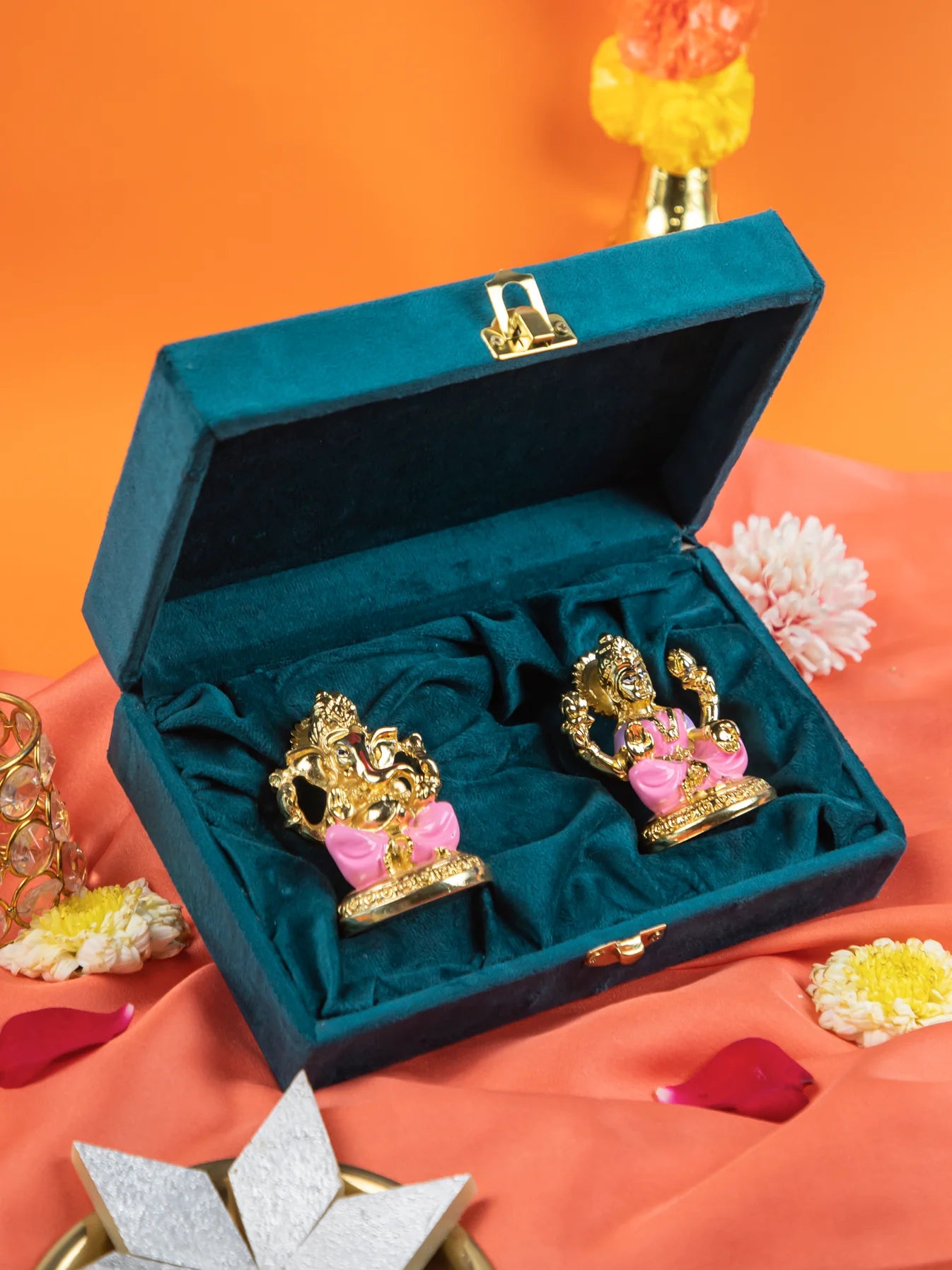 An image of an open teal blue velvet box containing two gold-colored idols of the Hindu deities Lakshmi and Ganesha with pink accents. The box is set on a peach-colored cloth, with yellow and white flowers and petals scattered around it.