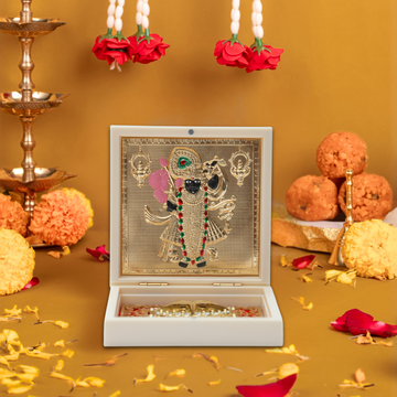 A beautifully arranged image featuring a gold-plated Shreenathji idol framed in an ornate white box, surrounded by traditional Hindu offerings like sweets (ladoo), marigold and rose petals, and a golden oil lamp stand, all set on a warm, golden-yellow background, evoking a sense of reverence and celebration.