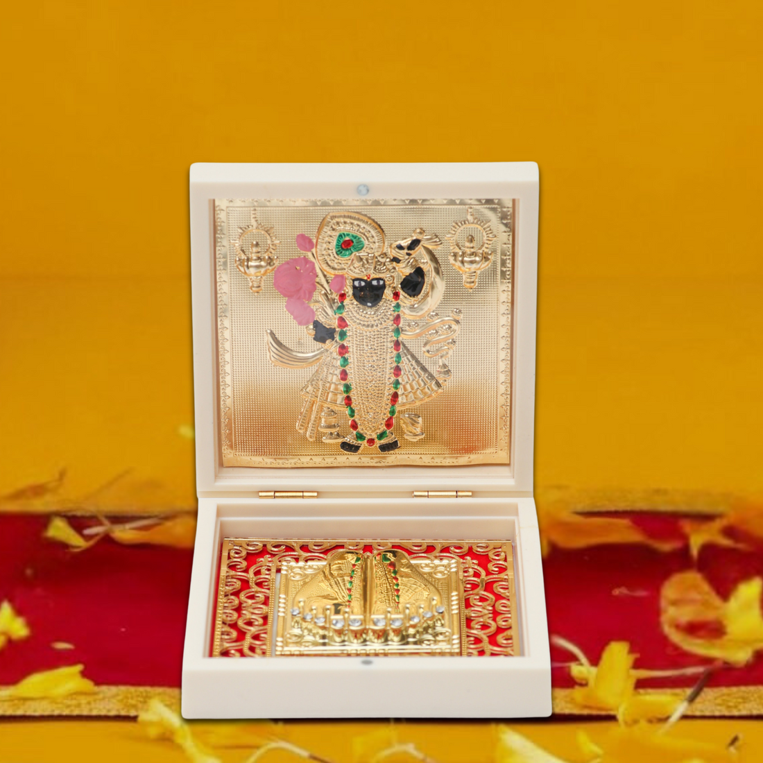 A beautifully arranged image featuring a gold-plated Shreenathji idol framed in an ornate white box, surrounded by traditional Hindu offerings like sweets (ladoo), marigold and rose petals, and a golden oil lamp stand, all set on a warm, golden-yellow background, evoking a sense of reverence and celebration.