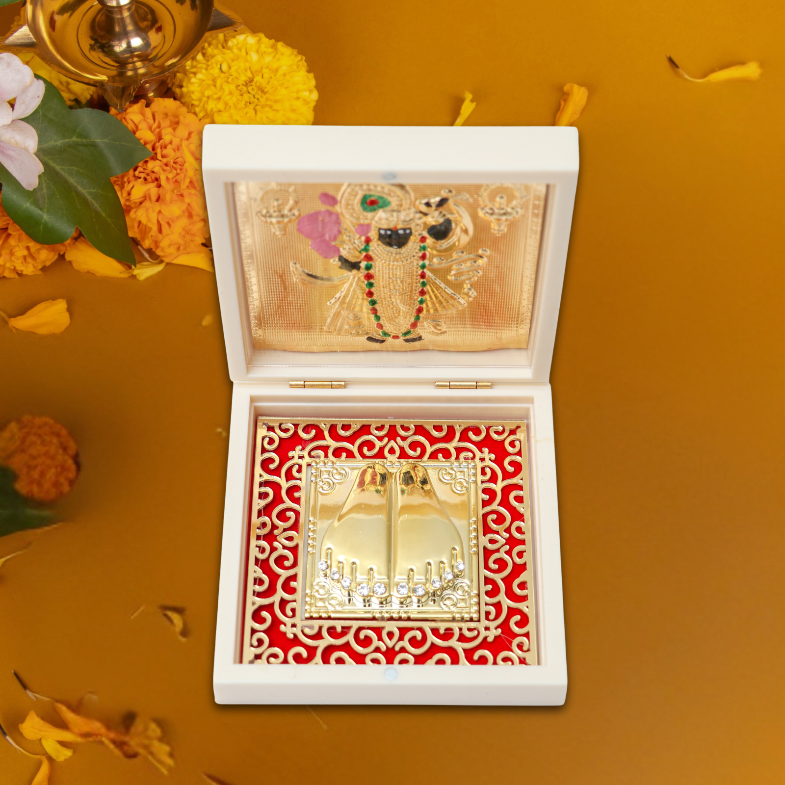 A white wooden box with a golden, multi-colored Shreenathji photo frame on the open lid, and a red velvet-lined compartment in the base holding gold-plated Shreenathji Charan Paduka (footprints), all set against a warm, golden background with scattered marigold flowers and petals, suggesting a sacred or celebratory context.