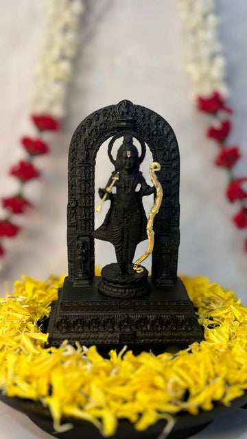 A black idol of Ram Lalla, depicting Lord Rama as a five-year-old child, standing with a golden bow and arrow, adorned with garlands of red and white flowers in the background and yellow flower petals scattered around the base.