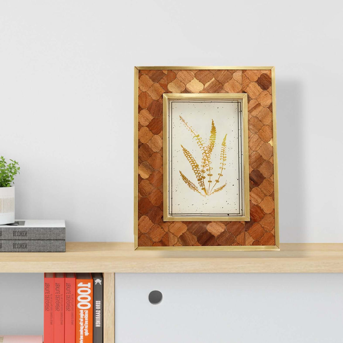 A teakwood frame with a geometric pattern, displaying a floral image, placed on a shelf with books and a plant in the background.