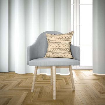 A cushion cover with a tribal geometric pattern on a chair in a room with wooden flooring and white curtains.