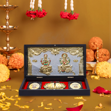 A detailed image showing a traditional Hindu puja set. The centerpiece is an open black display box with a red velvet interior, showcasing silver and gold-toned idols of Goddess Lakshmi and Lord Ganesha on a decorative backdrop. Below them are two gold-colored Laxmi Charan Paduka (footprints of Goddess Lakshmi) and a gold coin, symbolizing wealth and blessings. In the background, out of focus, are golden lamps, flower garlands, and sweets, suggesting a festive or devotional setting like Diwali celebrations.
