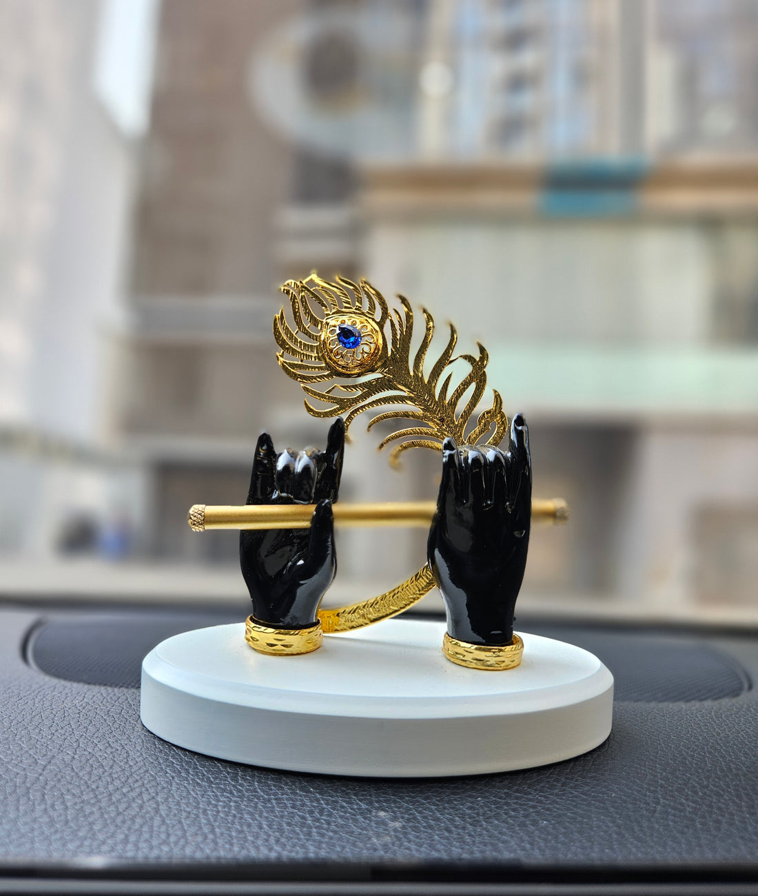 A decorative showpiece featuring two white sculpted hands, representing Lord Krishna's hands, gracefully holding a golden flute. A golden peacock feather with a blue jewel accent is positioned above the hands. The sculpture rests on a white, round base.