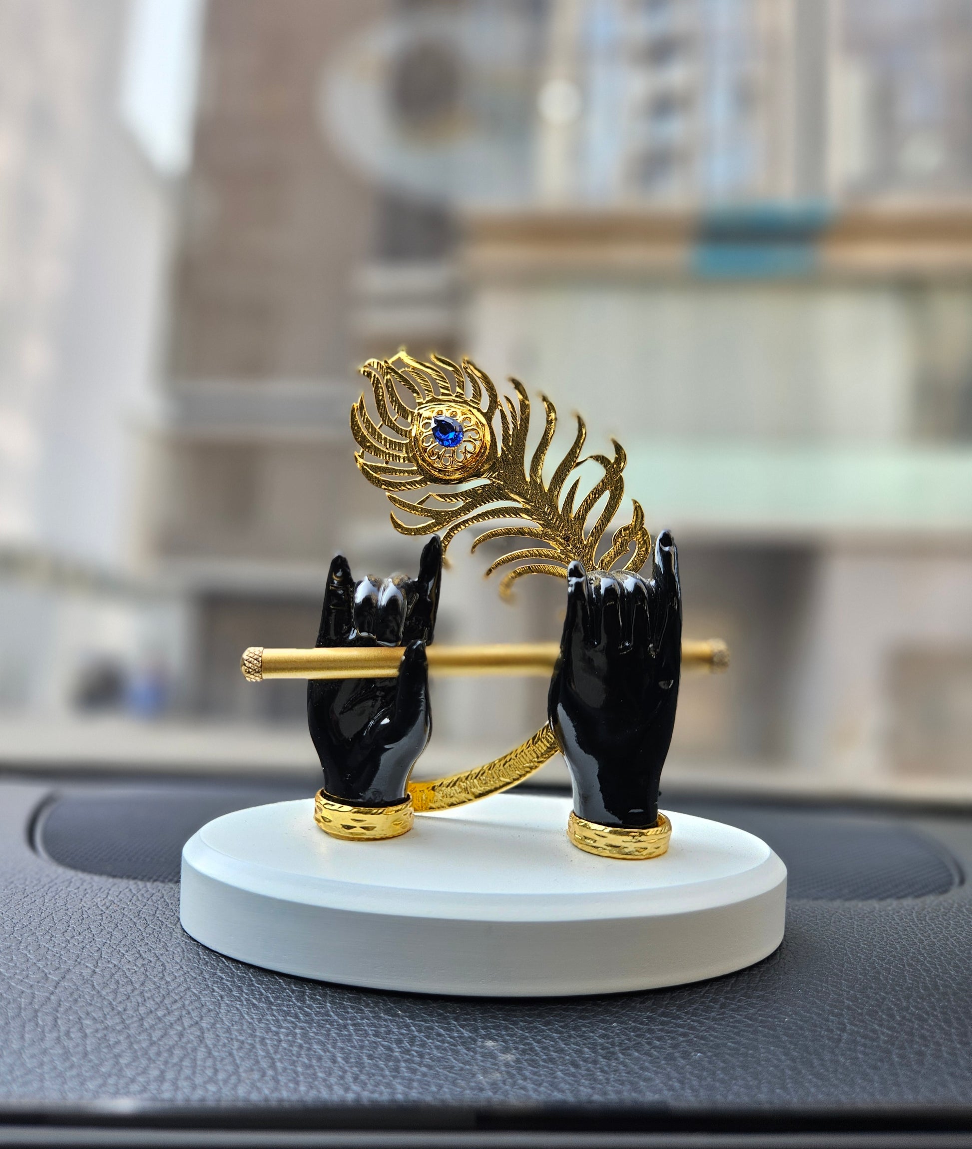 A decorative idol of Lord Krishna's black hands gracefully holding a golden flute, with a golden peacock feather featuring a blue eye detail rising from behind the flute, all mounted on a white circular base and placed on a car dashboard.