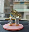 A gold-plated Lord Krishna hands idol holding a flute, adorned with a peacock feather, and a small Krishna face, set on a pink oval base, likely intended for car dashboard or home decor.
