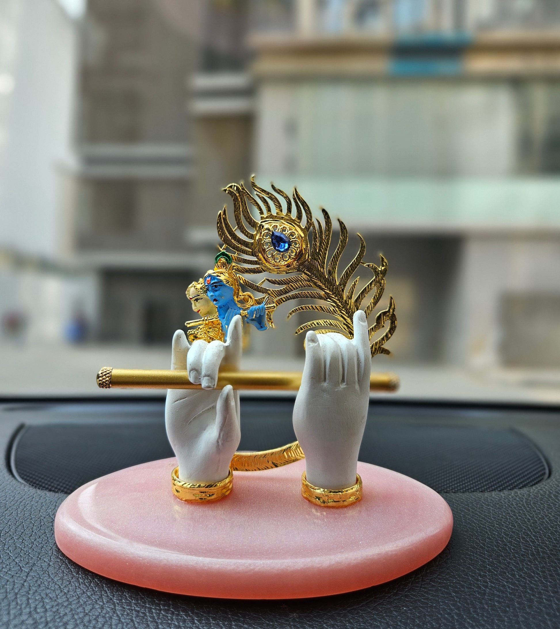 A gold-plated Lord Krishna hands idol holding a flute, adorned with a peacock feather, and a small Krishna face, set on a pink oval base, likely intended for car dashboard or home decor.