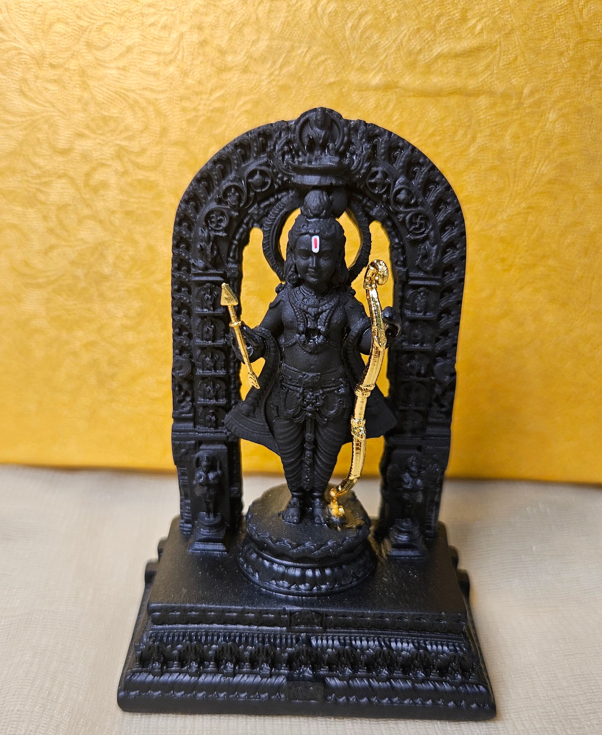 A black statue of a deity, likely Lord Rama, holding a golden bow and arrow is shown on a decorative base. The statue is set against a yellow, textured background and is framed by an intricate black archway.