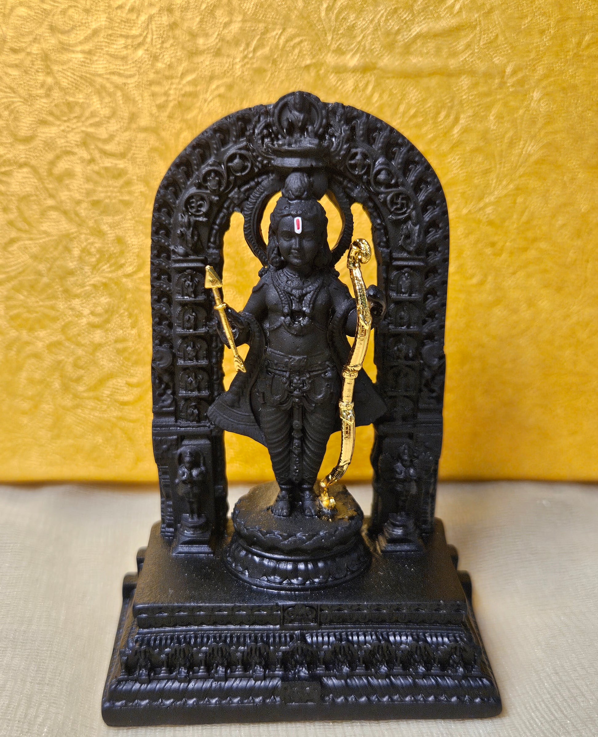 A black statue of a deity, likely Lord Rama, holding a golden bow and arrow is shown on a decorative base. The statue is set against a yellow, textured background and is framed by an intricate black archway.