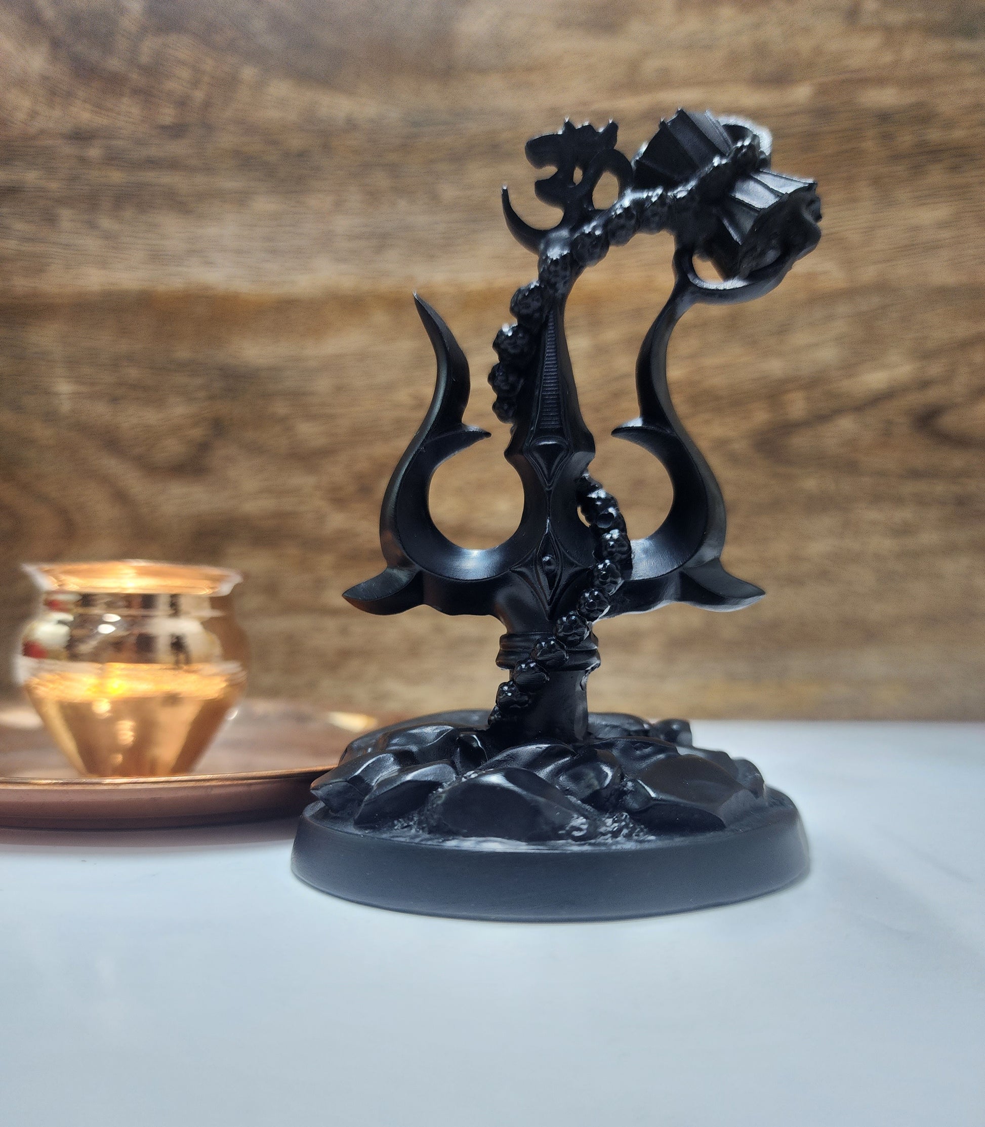 A black resin statue of Lord Shiva's Trishul (trident) with an attached Damru (drum) and an Om symbol, sitting on a base, likely a home decor showpiece
