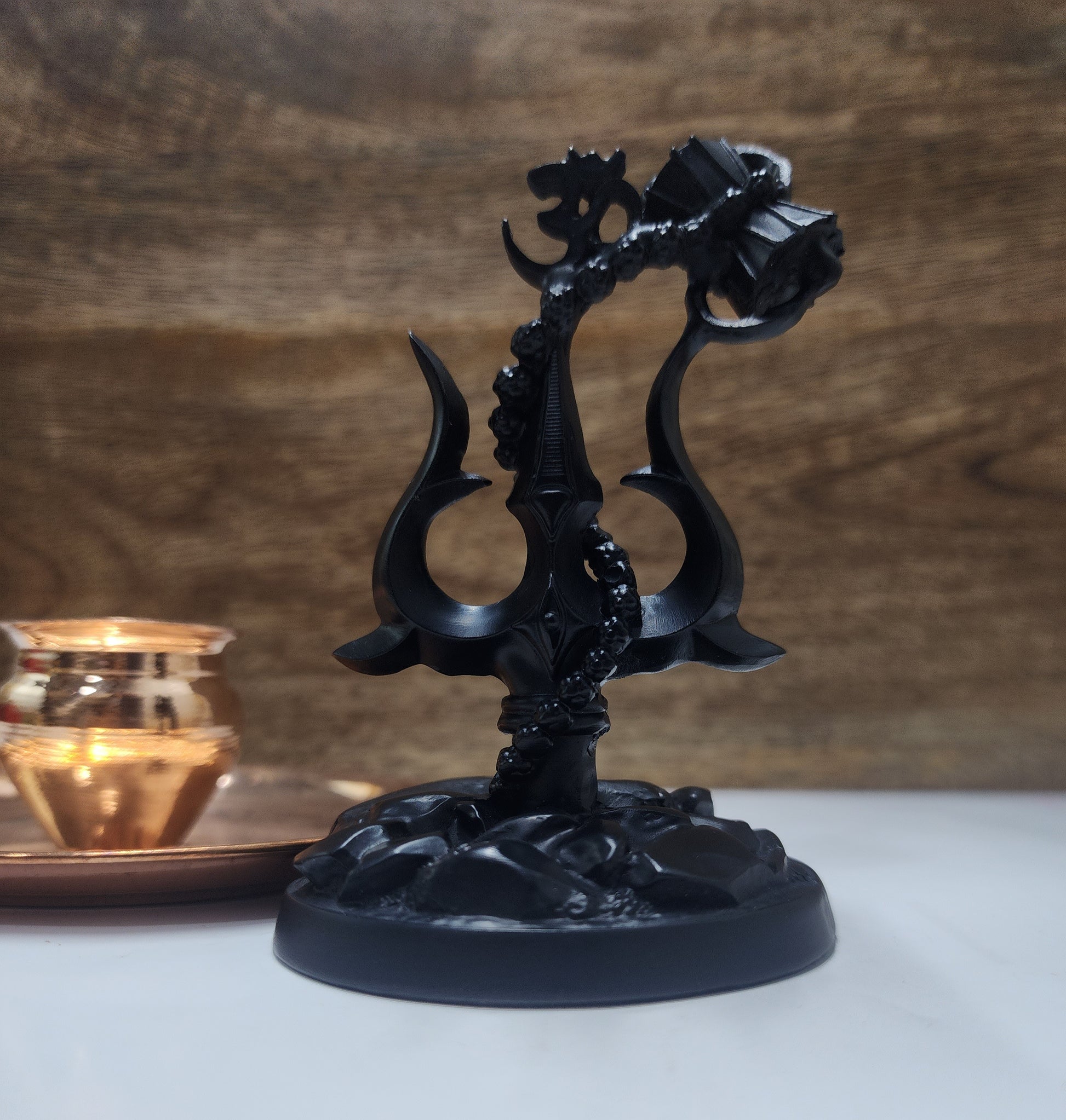 A black statue featuring a Shiva Trishul with a Damaru (drum) attached, and an Om symbol at its peak, all set on a dark, textured base.