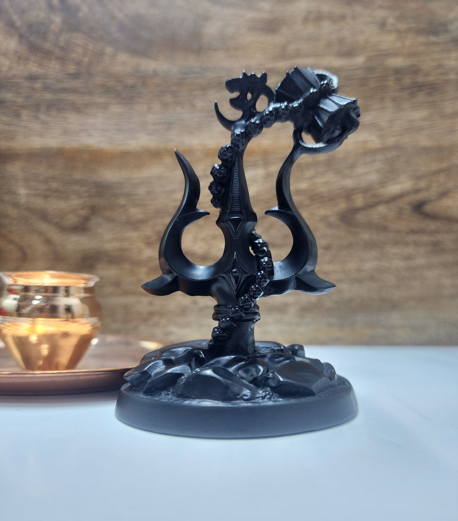 A black religious showpiece featuring a Shiva Trishul with an attached Damru drum and an Om symbol, standing on a textured base, with a blurry background showing a wooden surface and a warm glowing light source like a lamp or candle.