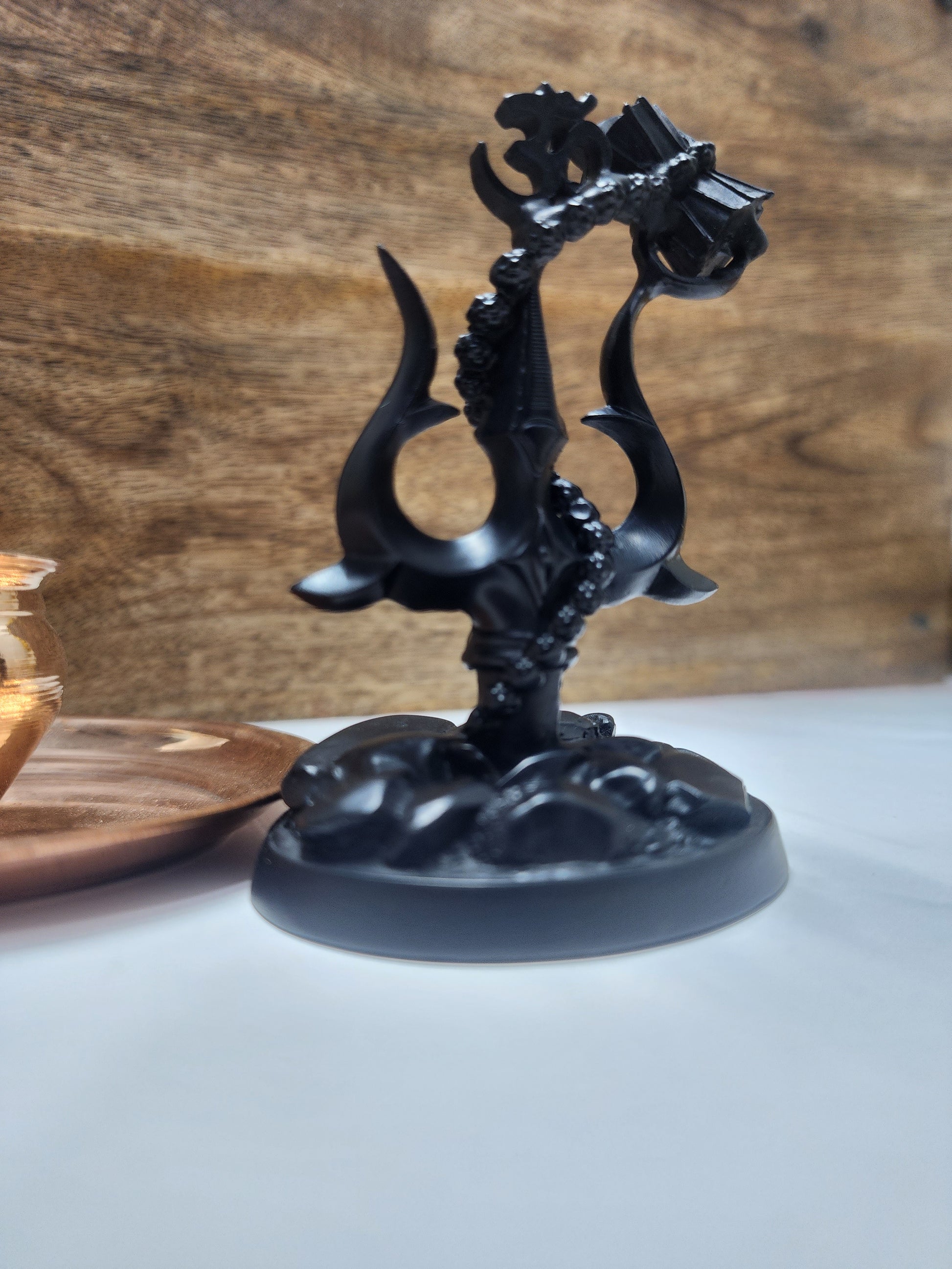 A black matte finished figurine of a Lord Shiva's Trishul (trident) with an attached Damru (drum) on a round base.