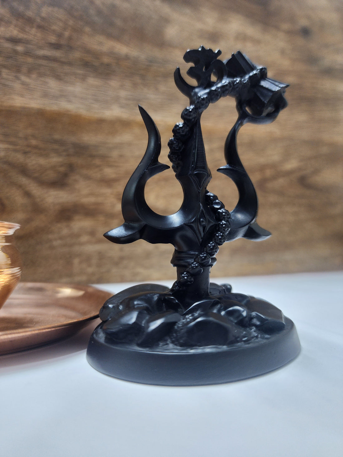 A black statue featuring a Shiva Trishul with a Damaru (drum) attached, and an Om symbol at its peak, all set on a dark, textured base.