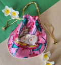 A pink satin potli bag featuring a printed illustration of a white Vastu Kamdhenu cow surrounded by pink lotus flowers, adorned with a beaded handle and pearl trim, displayed against a dual-colored green and beige background with white frangipani flowers.