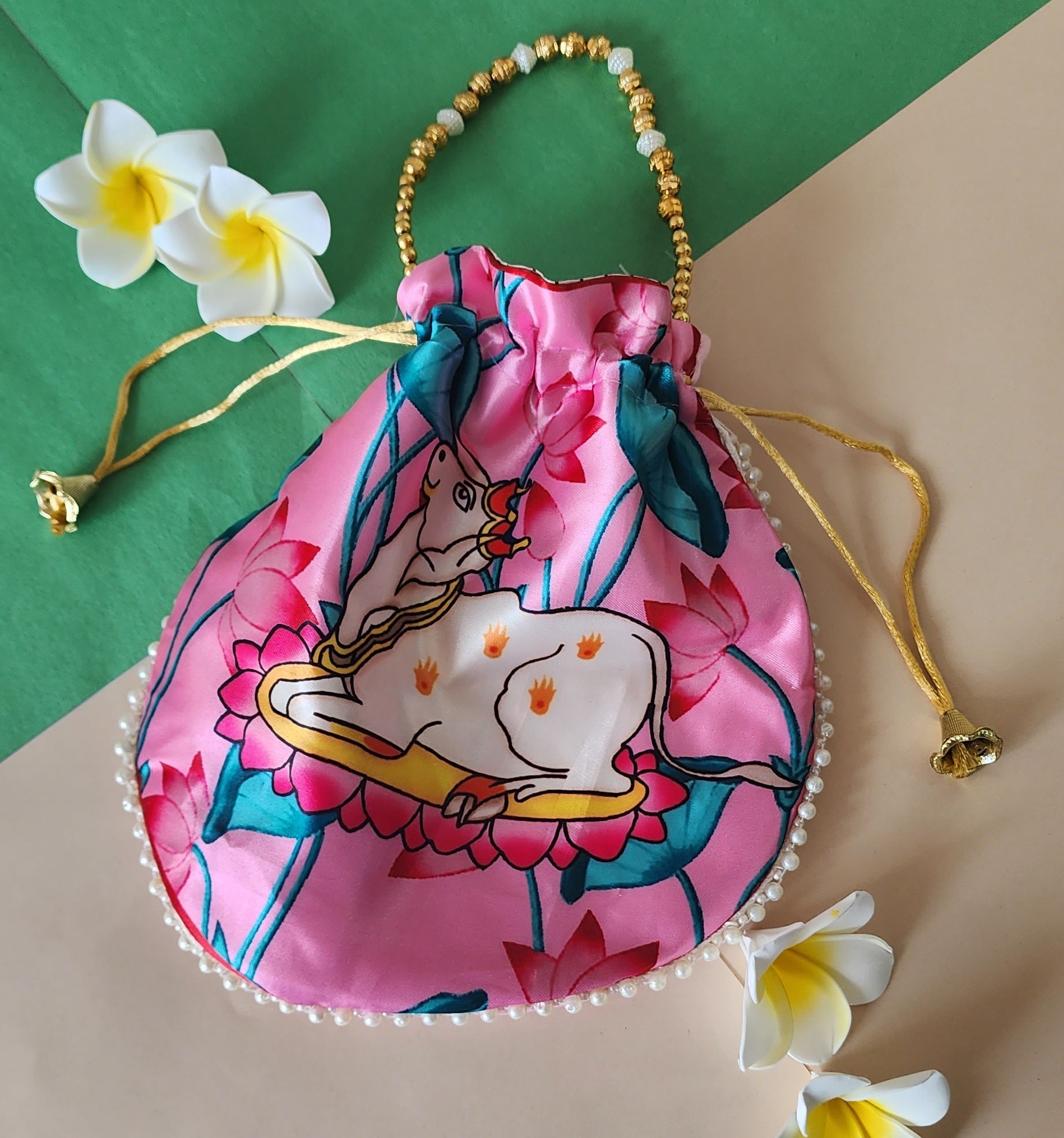 A pink satin potli bag featuring a printed illustration of a white Vastu Kamdhenu cow surrounded by pink lotus flowers, adorned with a beaded handle and pearl trim, displayed against a dual-colored green and beige background with white frangipani flowers.