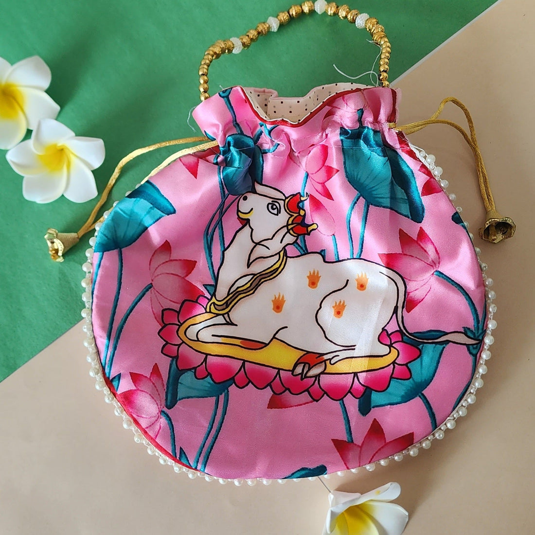 A vibrant pink Vastu Kamdhenu cow potli bag featuring a golden drawstring and handle, adorned with a white cow and calf illustration surrounded by pink lotus flowers.