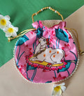 A pink Vastu Kamdhenu Cow Potli Bag featuring a sacred cow illustration surrounded by pink lotus flowers, with a golden beaded handle and pearl trim along the base.