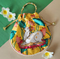 A yellow satin potli bag featuring a colorful print of a white Kamdhenu cow resting on a pink lotus flower, surrounded by green lotus leaves and pink lotus buds. The bag has a beaded gold handle and a decorative pearl trim along the lower edge.
