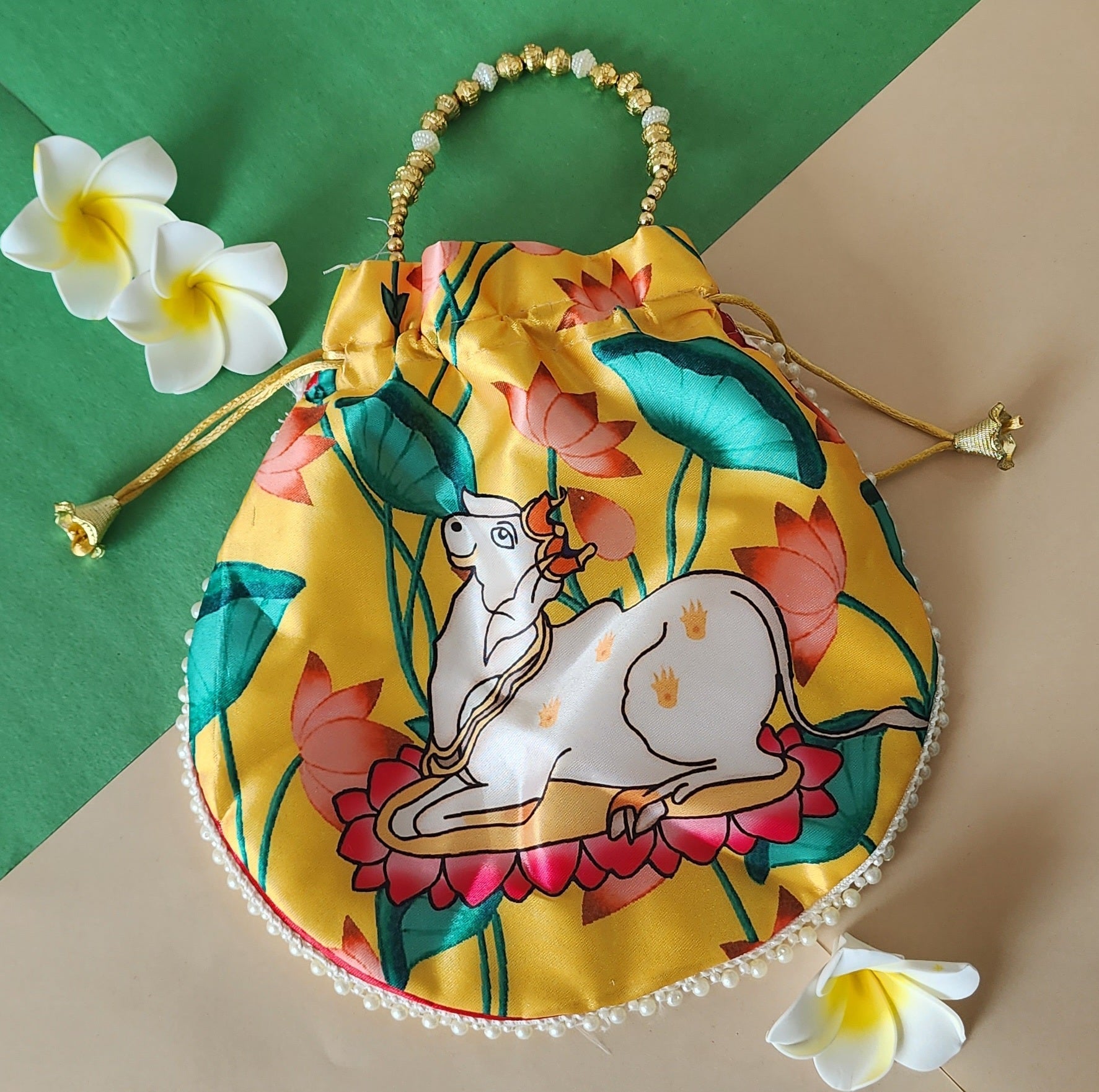 A yellow satin potli bag featuring a colorful print of a white Kamdhenu cow resting on a pink lotus flower, surrounded by green lotus leaves and pink lotus buds. The bag has a beaded gold handle and a decorative pearl trim along the lower edge.