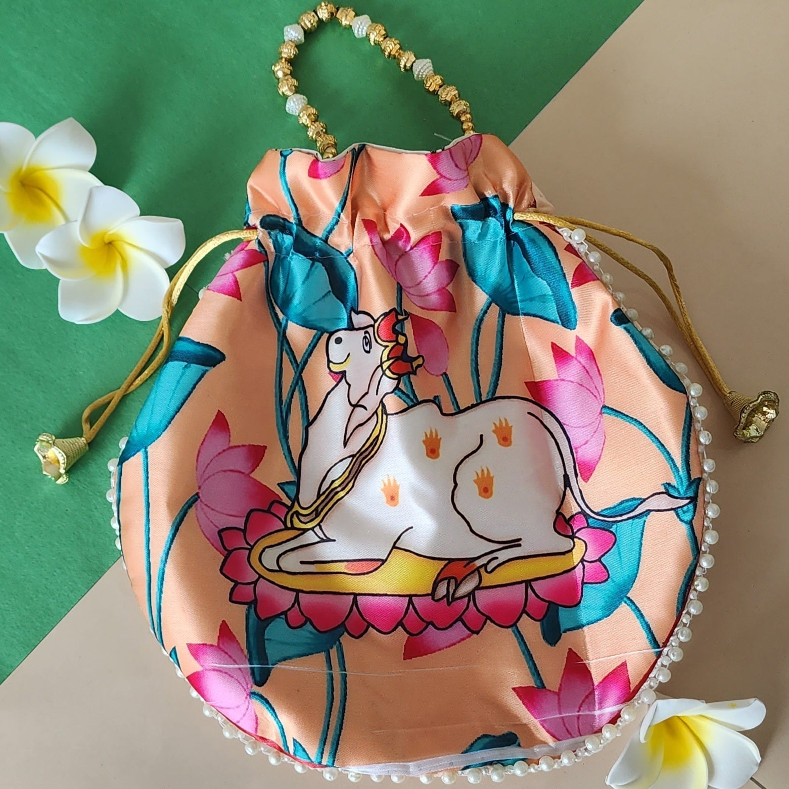 A peach-colored potli bag featuring a printed illustration of a white Kamdhenu cow surrounded by pink lotus flowers and green leaves, with a beaded handle and pearl embellishments along the bottom edge.