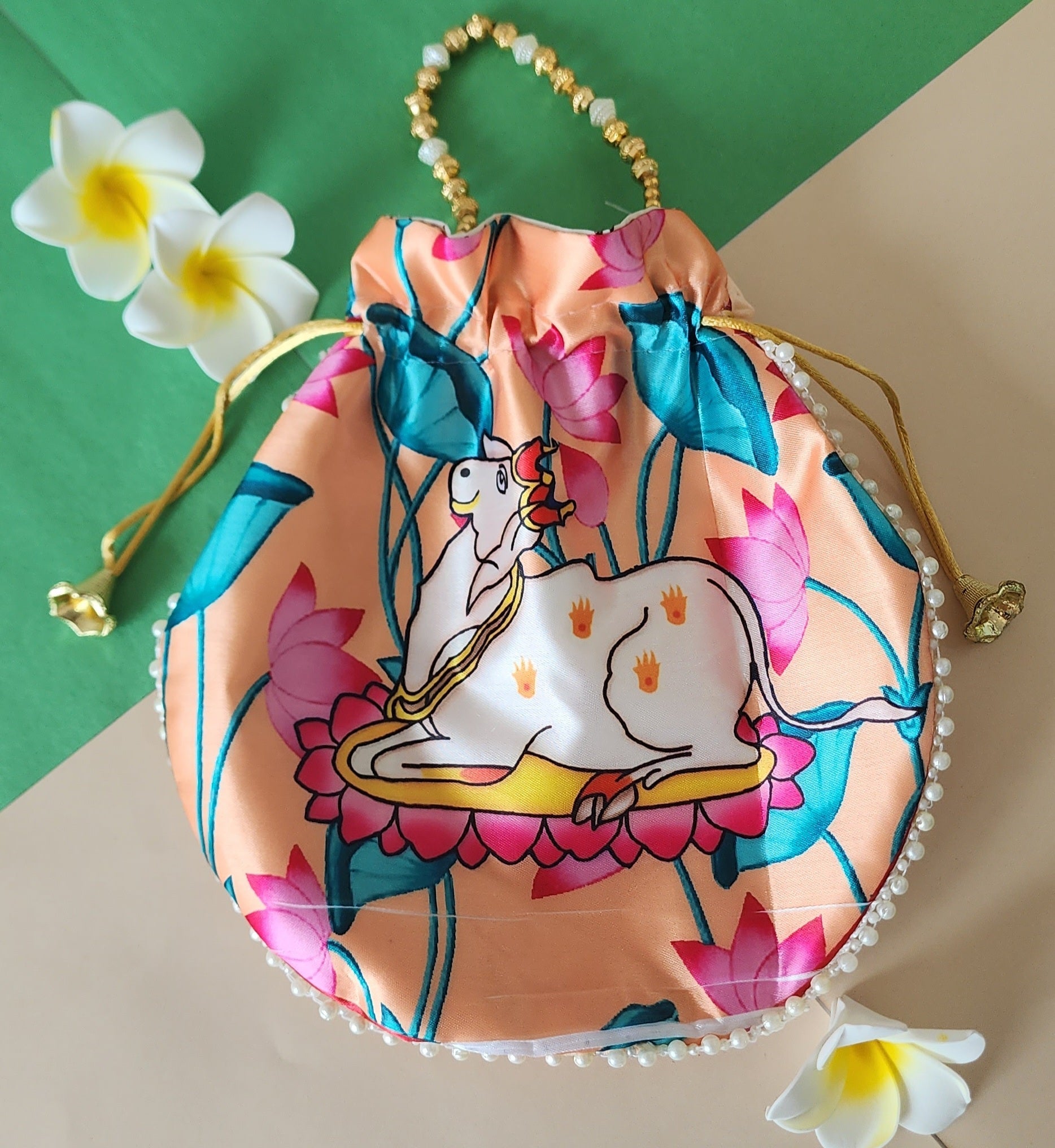 A Vastu Kamdhenu Cow Potli Bag in a peach color, featuring a printed illustration of the sacred Kamdhenu cow with lotus motifs, and embellished with pearl details along the bottom edge and a beaded handle.