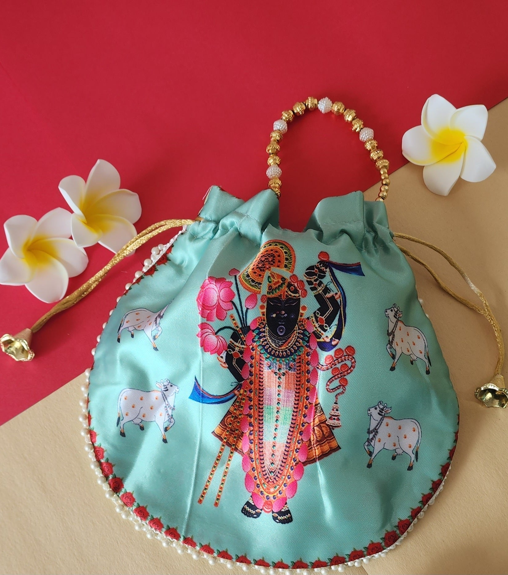 A light green Shreenathji Potli Bag featuring a colorful print of Lord Shreenathji, accented with a gold and white beaded handle and gold drawstring tassels. The bag rests on a red and beige surface, flanked by white frangipani flowers.