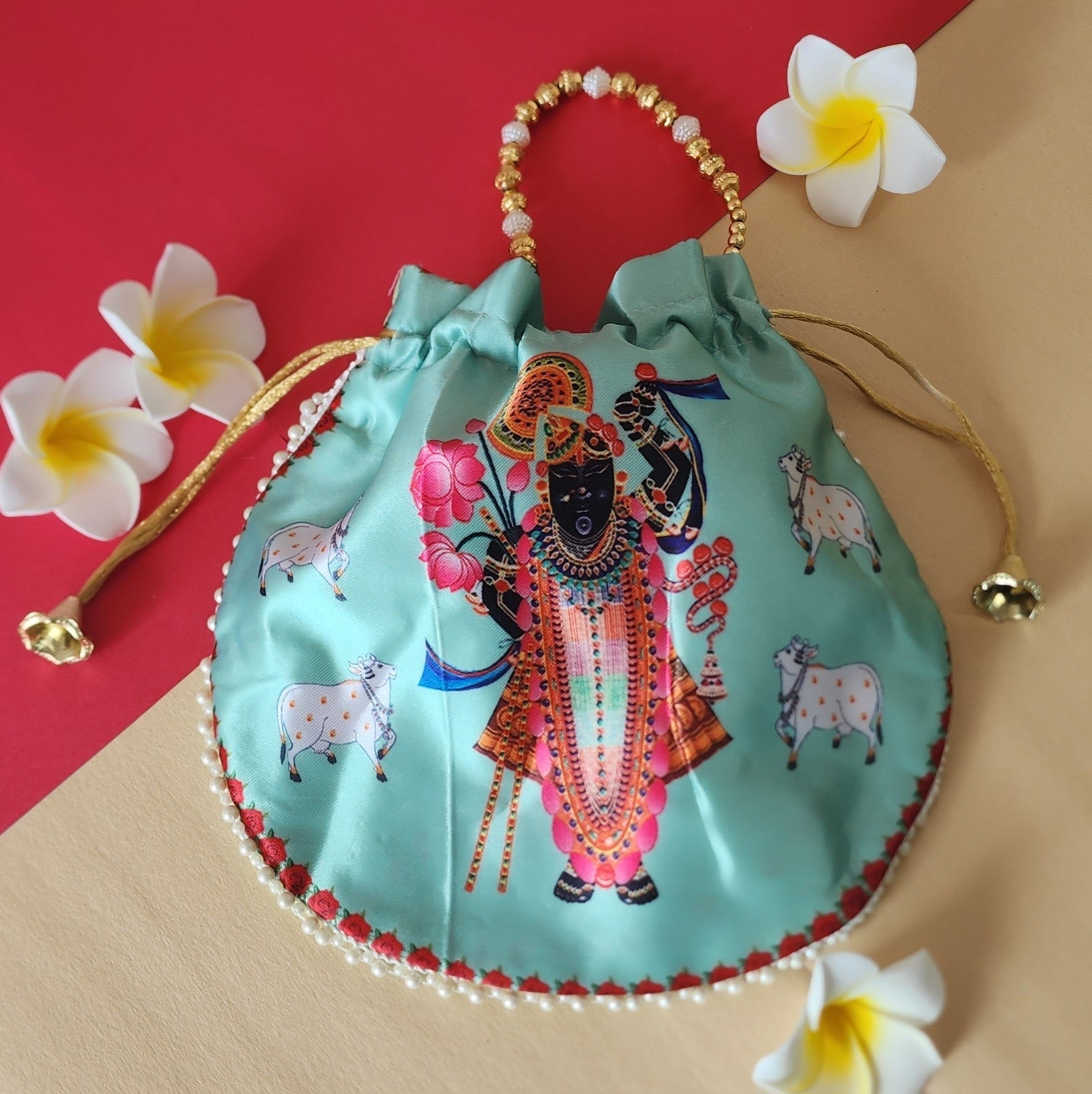 A beautiful, light teal/green satin potli bag (drawstring purse) with a digital print featuring a detailed image of Lord Shreenathji at the center, surrounded by white cows. The bag has a pearl-beaded handle and decorative golden drawstrings with tassels, resting on a red and beige background with white frangipani flowers arranged around it, enhancing its traditional and festive aesthetic