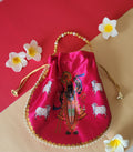 A vibrant pink Shreenathji Potli Bag featuring a divine Shreenathji print and cow motifs, adorned with pearl and gold beaded trim and handle, placed on a dual-toned red and beige background with white frangipani flowers.