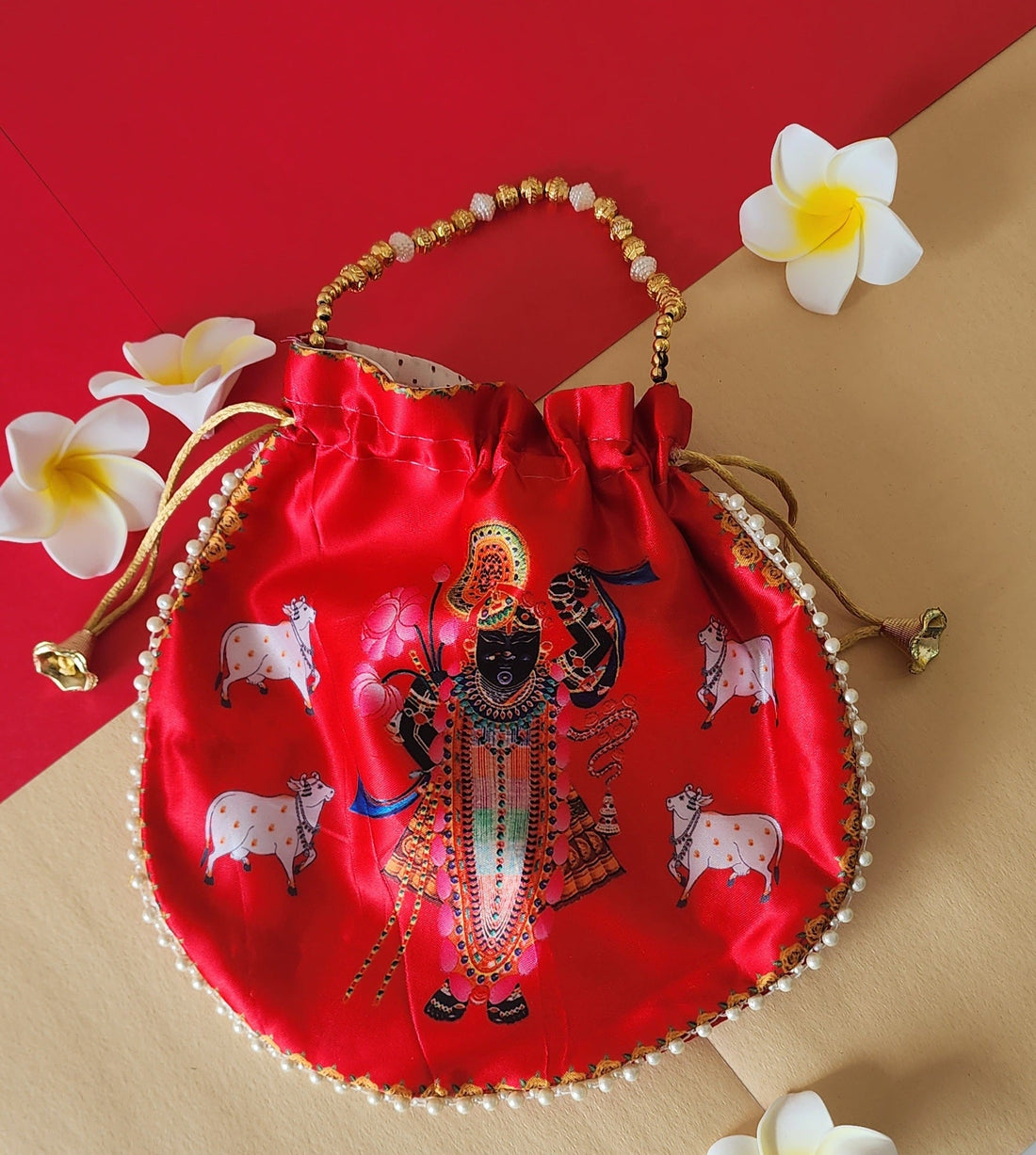 A light green Shreenathji Potli Bag featuring a colorful print of Lord Shreenathji, accented with a gold and white beaded handle and gold drawstring tassels. The bag rests on a red and beige surface, flanked by white frangipani flowers.
