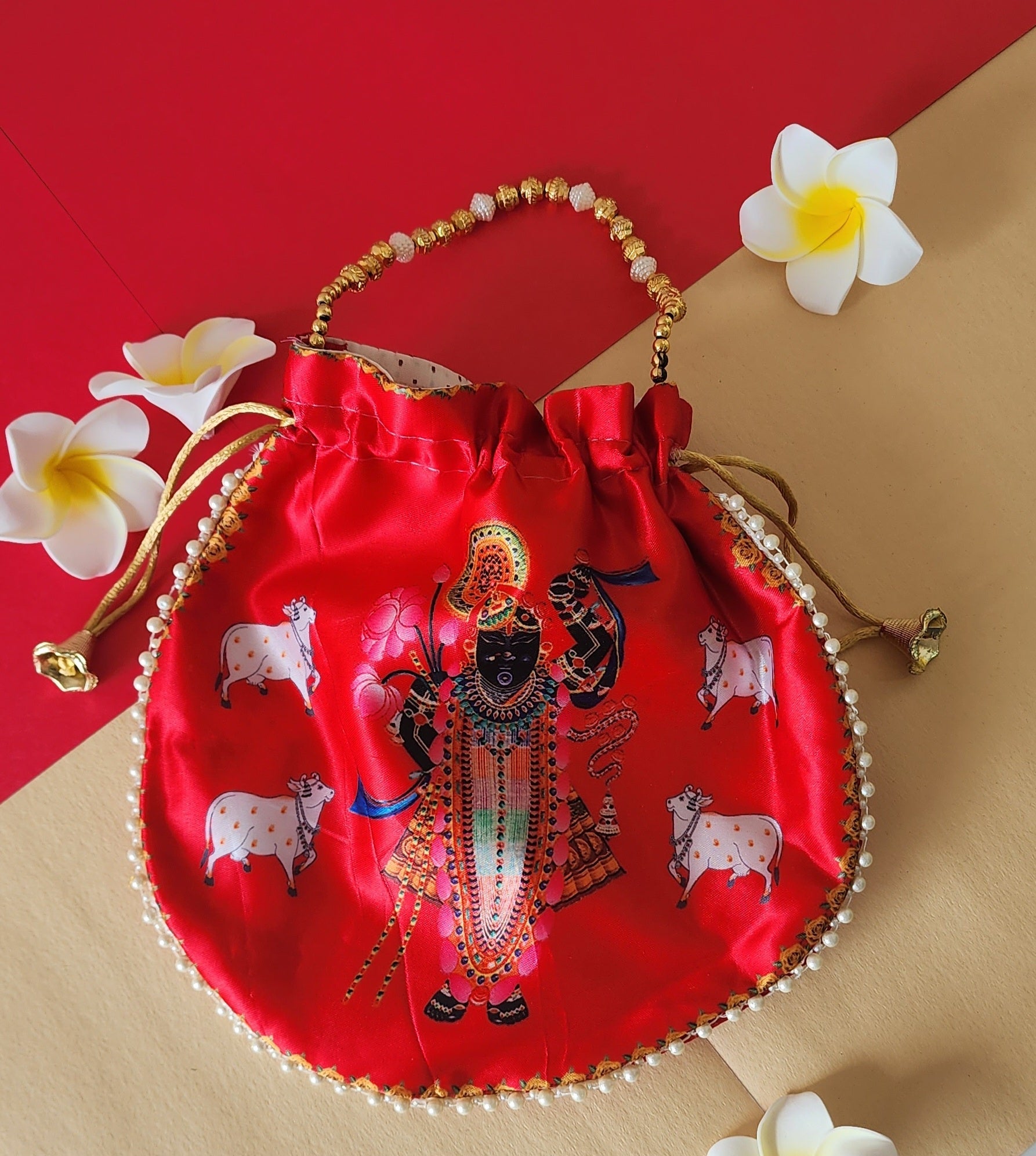 A vibrant red potli bag with a printed image of Shreenathji and cows, accented with a beaded handle and surrounded by white and yellow frangipani flowers, all set against a red and beige background.
