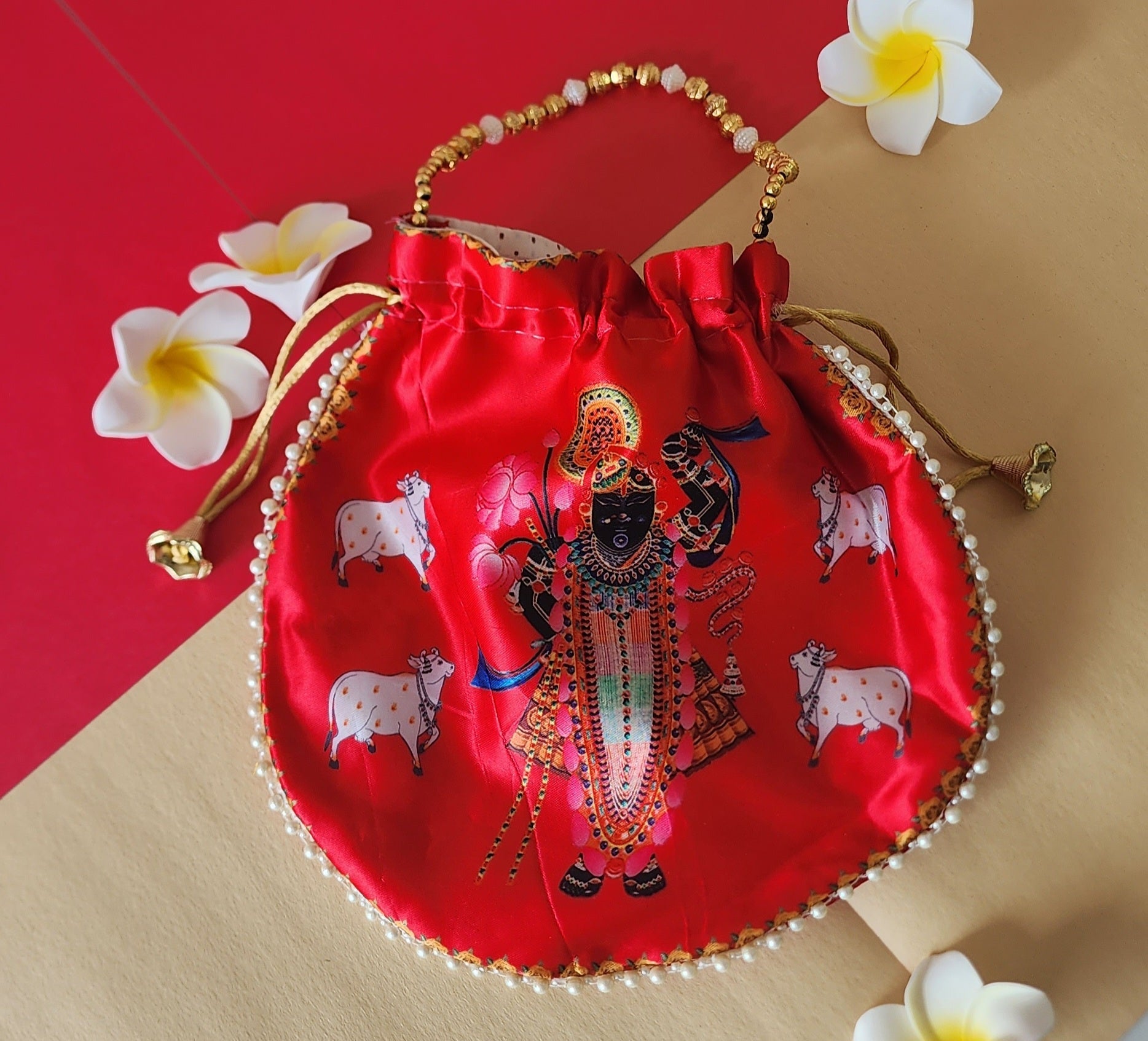 A vibrant red silk potli bag featuring a printed illustration of Shreenathji surrounded by cows, accented with pearl trim along the edge and a golden beaded handle.