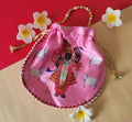 A detailed image of a vibrant pink Shreenathji Potli bag featuring a colorful print of Shreenathji (Lord Krishna) surrounded by cows. The traditional drawstring bag has a gold and pearl beaded handle and is embellished with small white pearls along its bottom edge. It is artfully arranged on a two-toned background, with several white frangipani flowers adding an elegant touch.