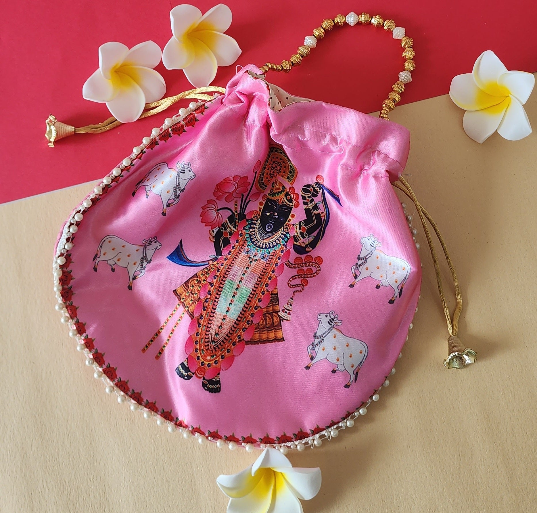 A vibrant pink Shreenathji potli bag with a detailed print of Lord Shreenathji and cows, featuring a pearl-beaded handle and gold drawstring with tassels, displayed on a contrasting red and beige background with white plumeria flowers.