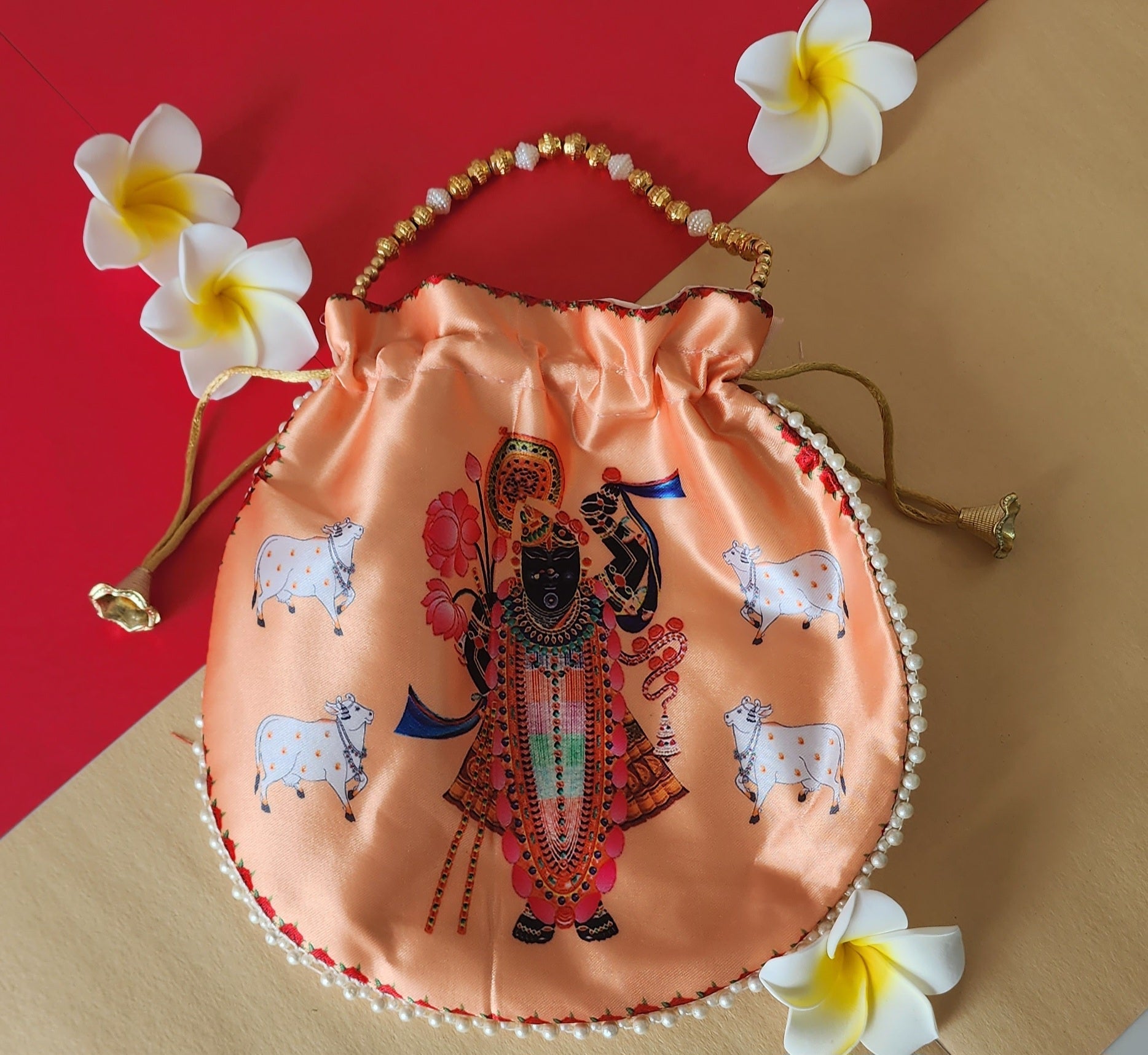 A peach-colored satin Potli bag featuring a digital print of Shreenathji with four cows, adorned with a white pearl and gold bead handle, and surrounded by white frangipani flowers against a red and beige background.