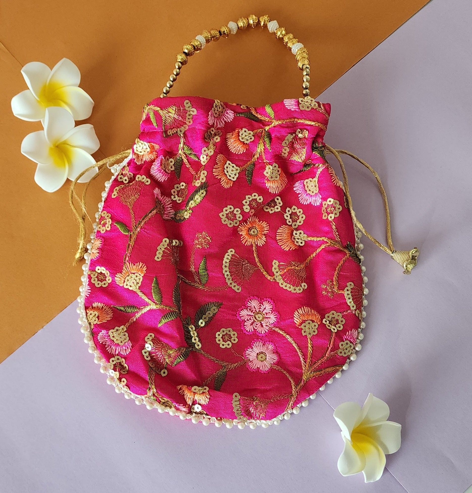 A pink floral design potli bag with golden embroidery and a drawstring closure, displayed alongside yellow flowers.