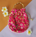A pink floral design potli bag with golden embroidery and a drawstring closure, displayed alongside yellow flowers.