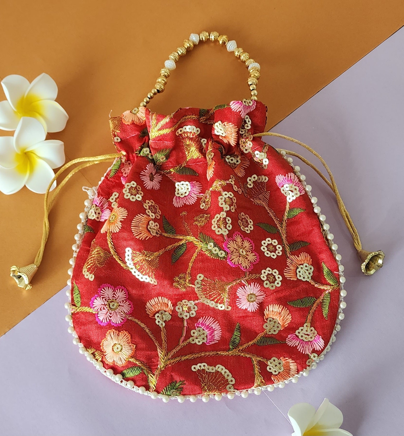A Orange floral design potli bag with golden embroidery and a drawstring closure, displayed alongside yellow flowers.