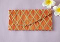 An orange envelope with a crisscross geometric pattern, displayed on a flat surface with a few flowers beside it.