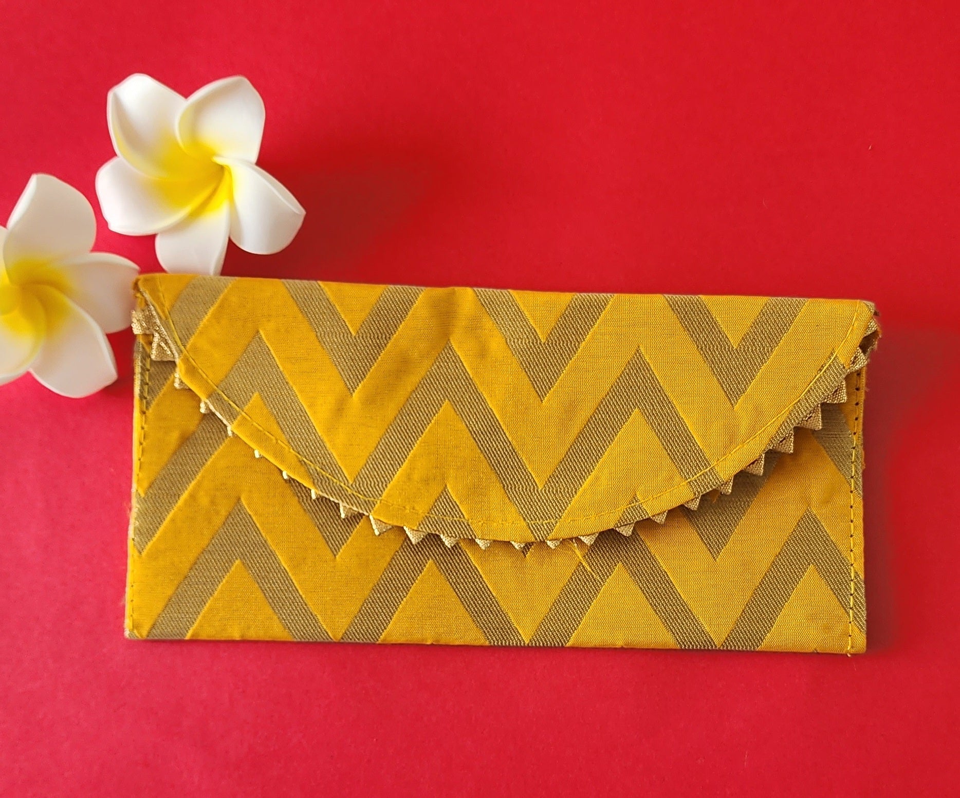 A V-shaped Yellow and gold shagun envelope with a zigzag pattern and decorative golden trimmings, displayed on an orange background with a frangipani flower beside it.