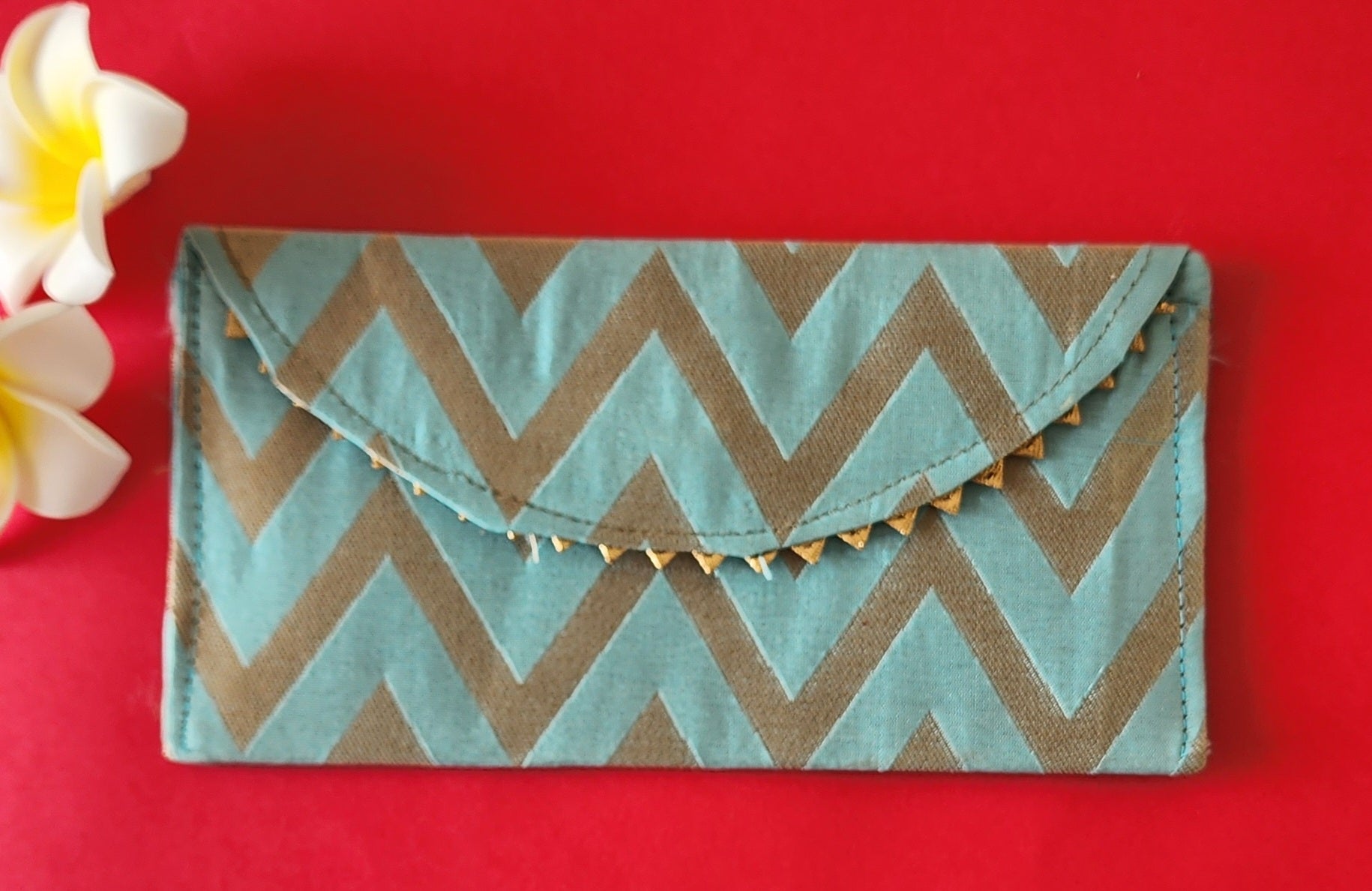 A V-shaped red and gold shagun envelope with a zigzag pattern and decorative golden trimmings, displayed on an orange background with a frangipani flower beside it.