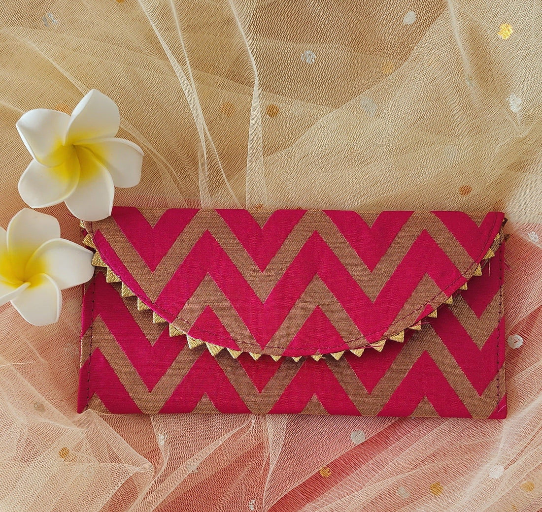 A V-shaped red and gold shagun envelope with a zigzag pattern and decorative golden trimmings, displayed on an orange background with a frangipani flower beside it.