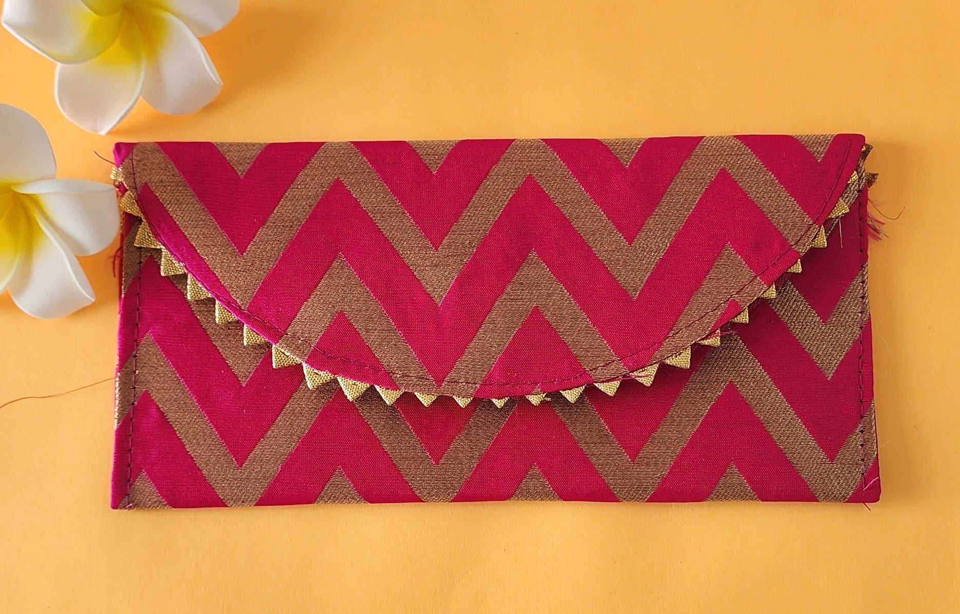 A V-shaped red and gold shagun envelope with a zigzag pattern and decorative golden trimmings, displayed on an orange background with a frangipani flower beside it.