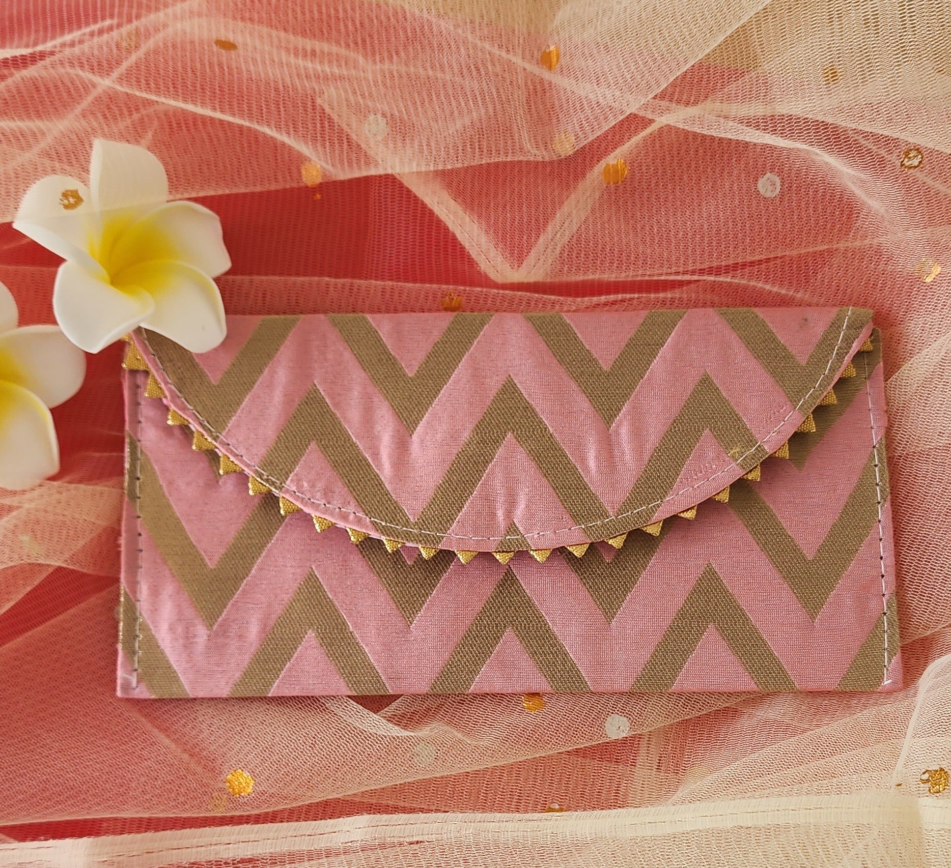 A V-shaped red and gold shagun envelope with a zigzag pattern and decorative golden trimmings, displayed on an orange background with a frangipani flower beside it.